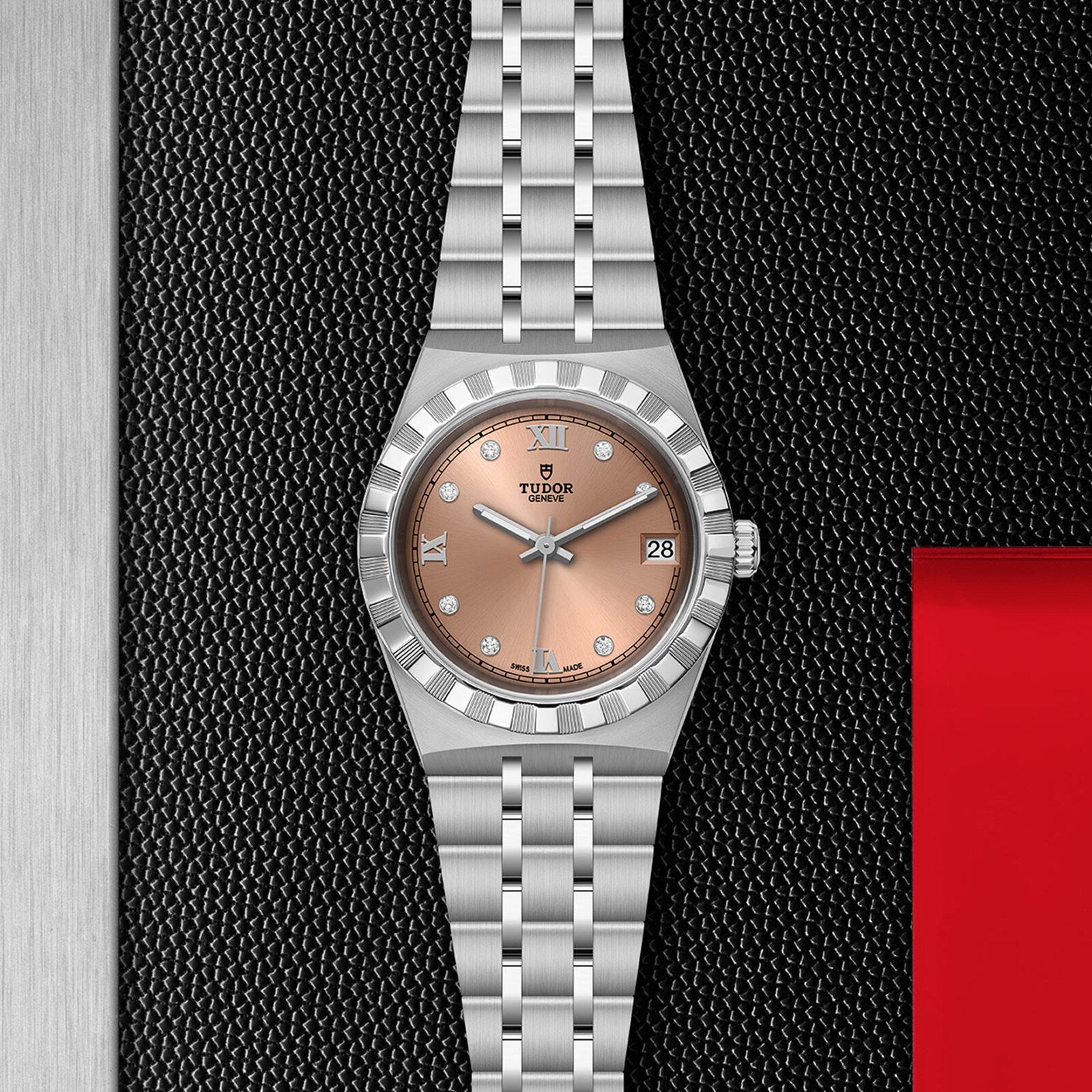 A TUDOR Royal stainless steel watch with a pink dial, diamond hour markers, automatic movement, and metal bracelet is shown on a textured black surface with metallic and red accents.