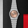 A TUDOR Royal stainless steel watch with a pink dial, diamond hour markers, automatic movement, and metal bracelet is shown on a textured black surface with metallic and red accents.