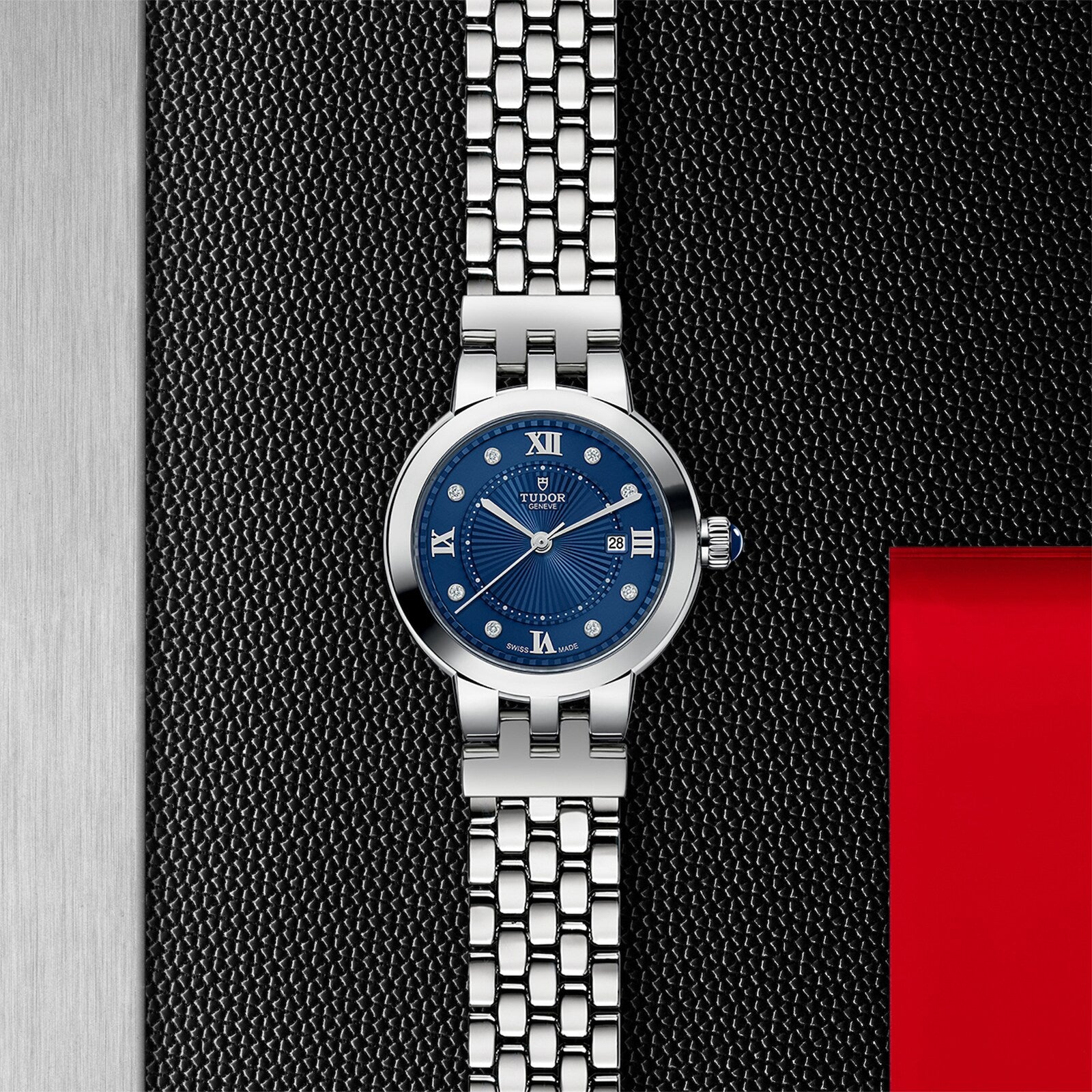 The TUDOR Clair de Rose watch features a blue dial, set against a textured black and silver background with a red accent on the right.