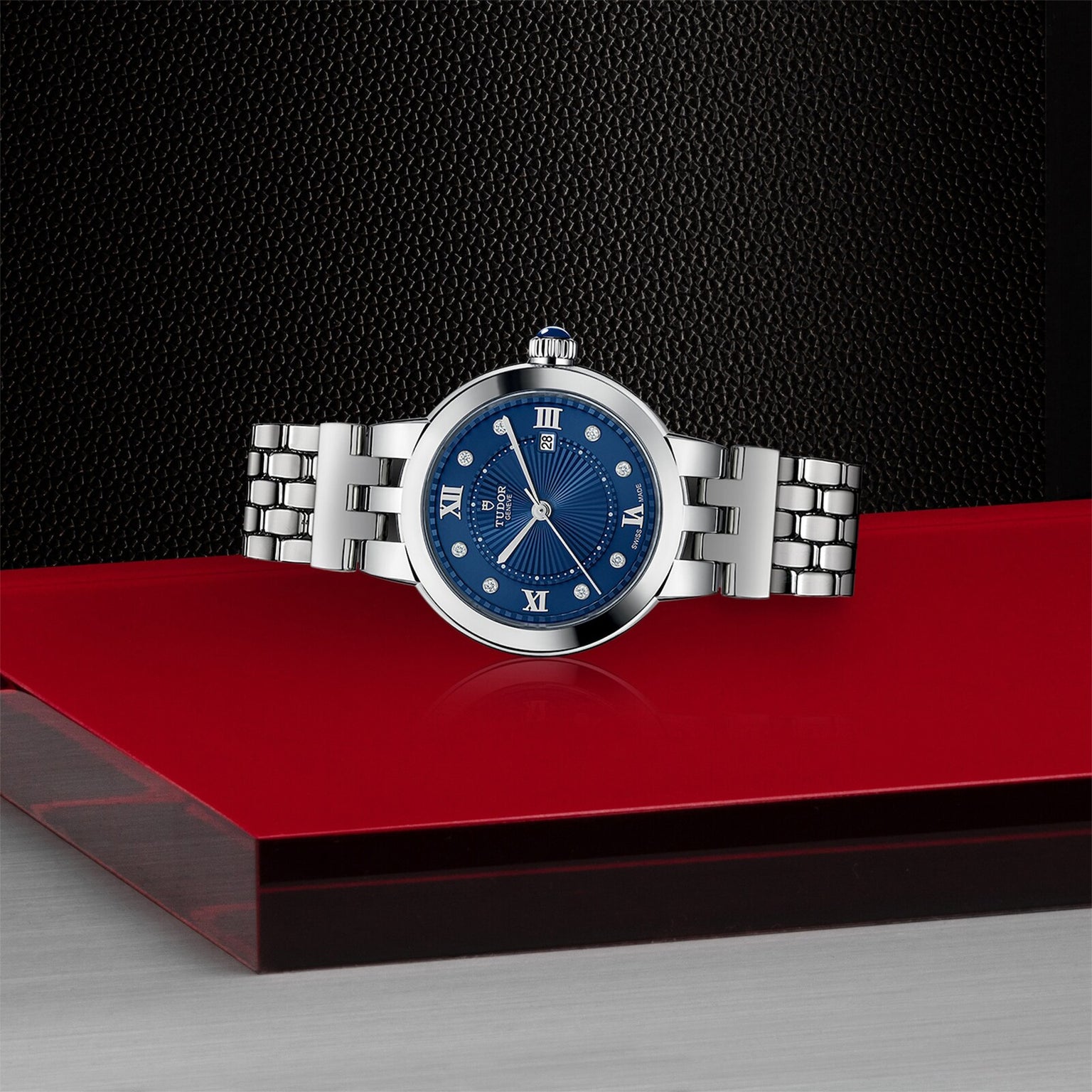 The TUDOR Clair de Rose is a silver wristwatch featuring a blue face and Roman numerals, displayed on a modern red and black surface.