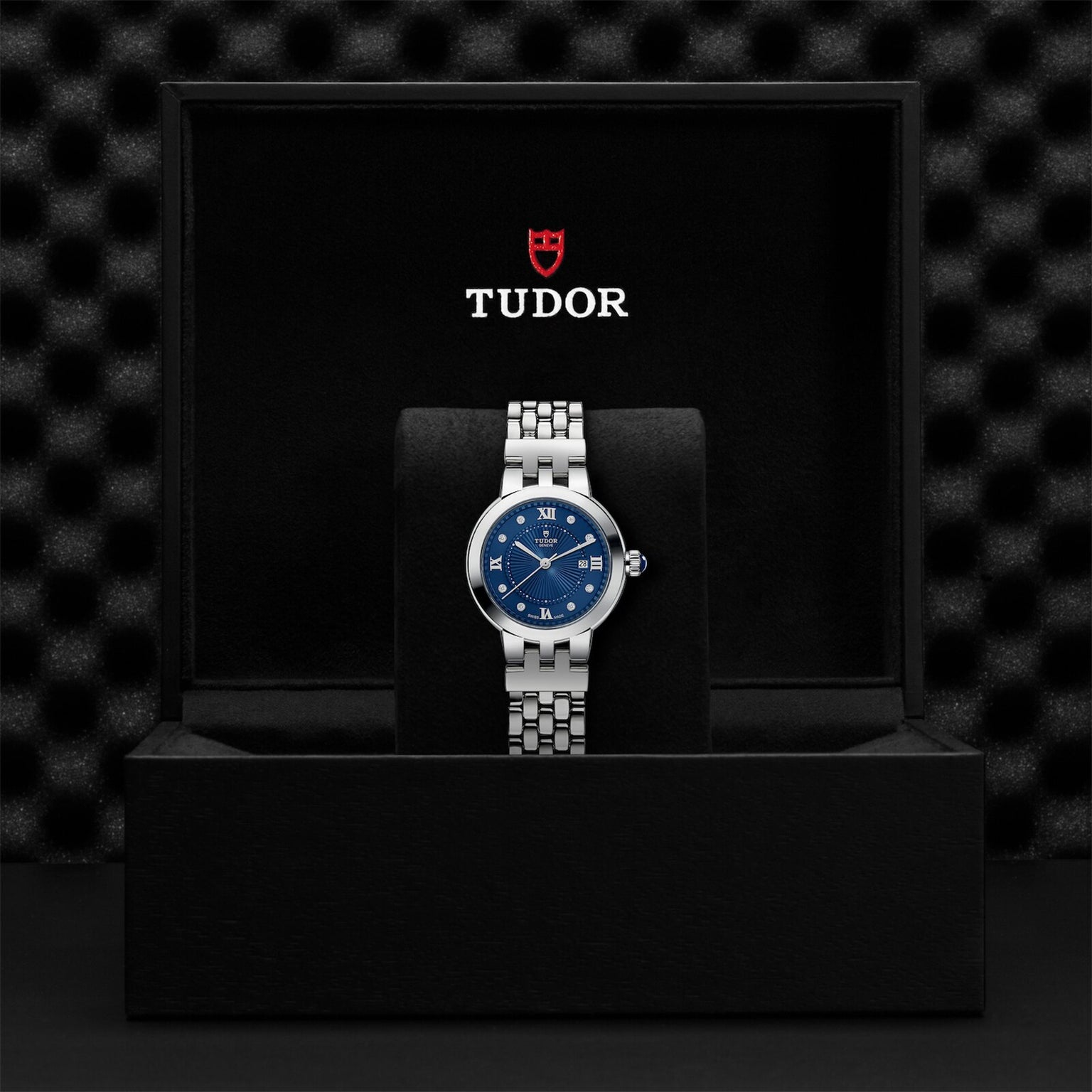 A TUDOR Clair de Rose wristwatch with a blue dial and silver band is displayed in an open black TUDOR box.