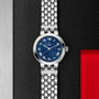 The TUDOR Clair de Rose watch with a blue dial and Roman numerals is displayed on a textured black and red background.