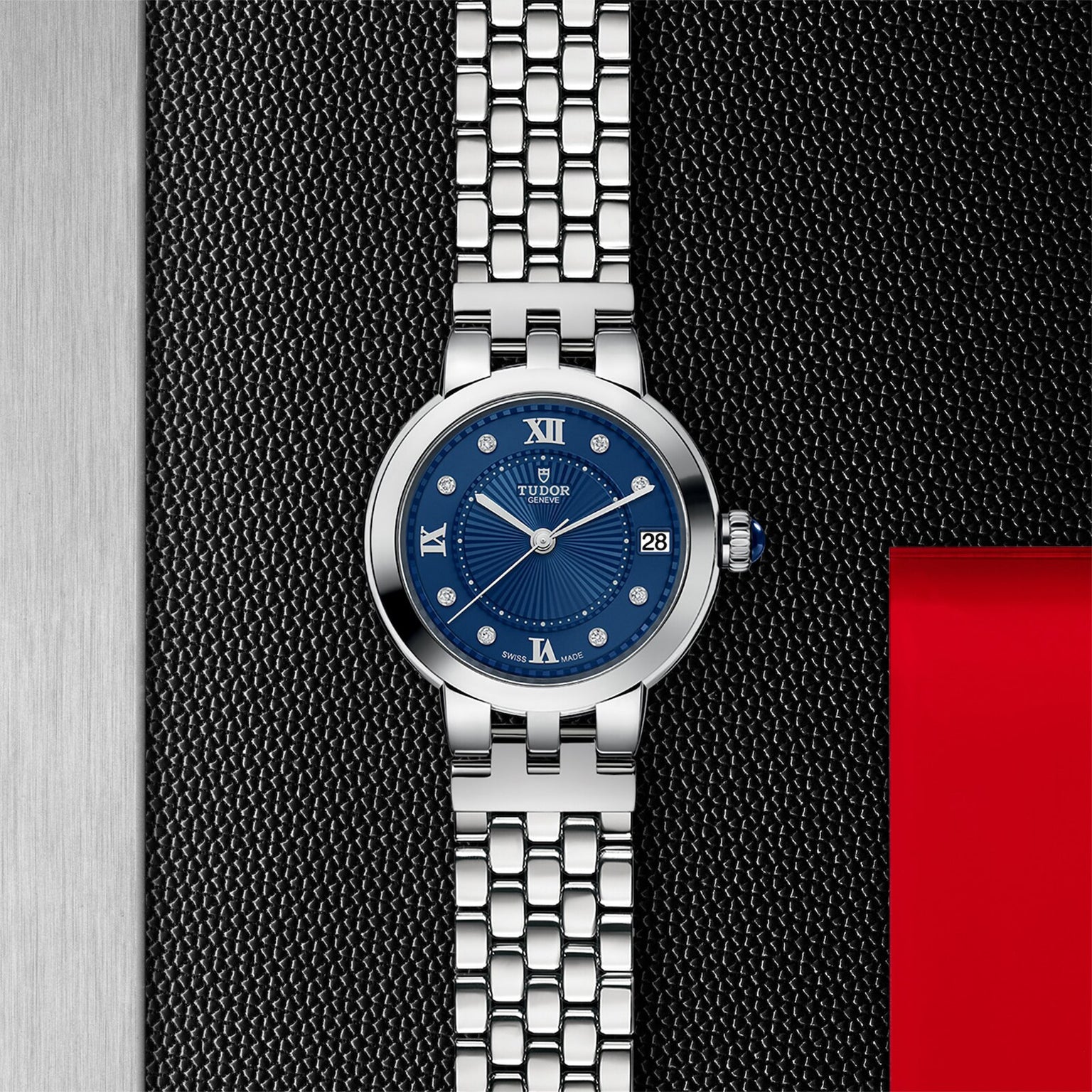 The TUDOR Clair de Rose wristwatch, featuring a blue dial and metal bracelet, is showcased on a textured black and metallic surface.