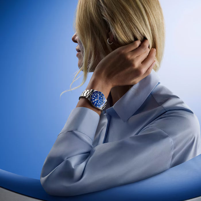 Person wearing light blue shirt with Black Bay 54 watch featuring blue dial and metal bracelet on left wrist.