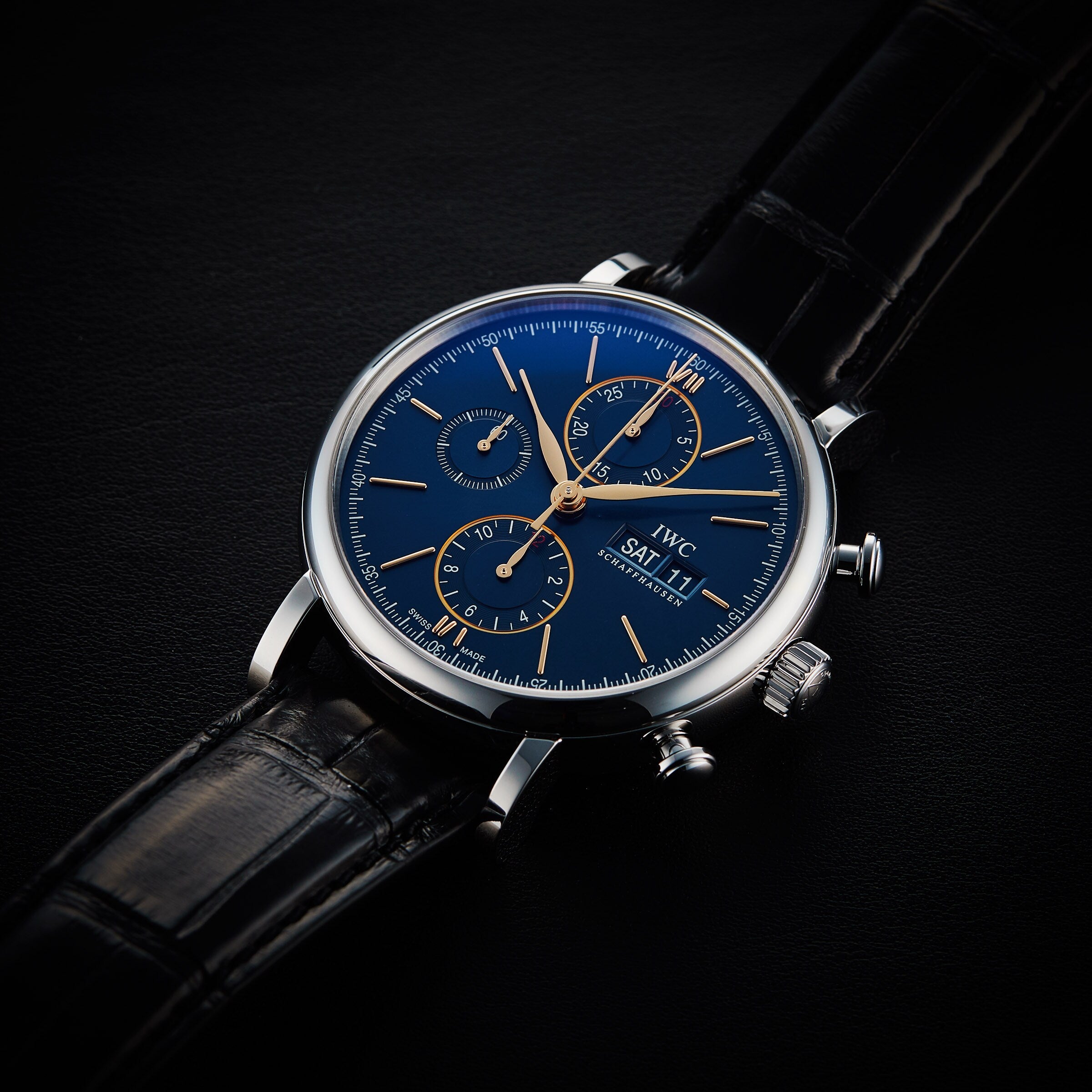 The IWC Schaffhausen Portofino Chronograph features a blue dial, gold hands and markers, three chronograph subdials, and a black leather strap, elegantly displayed on a dark surface.