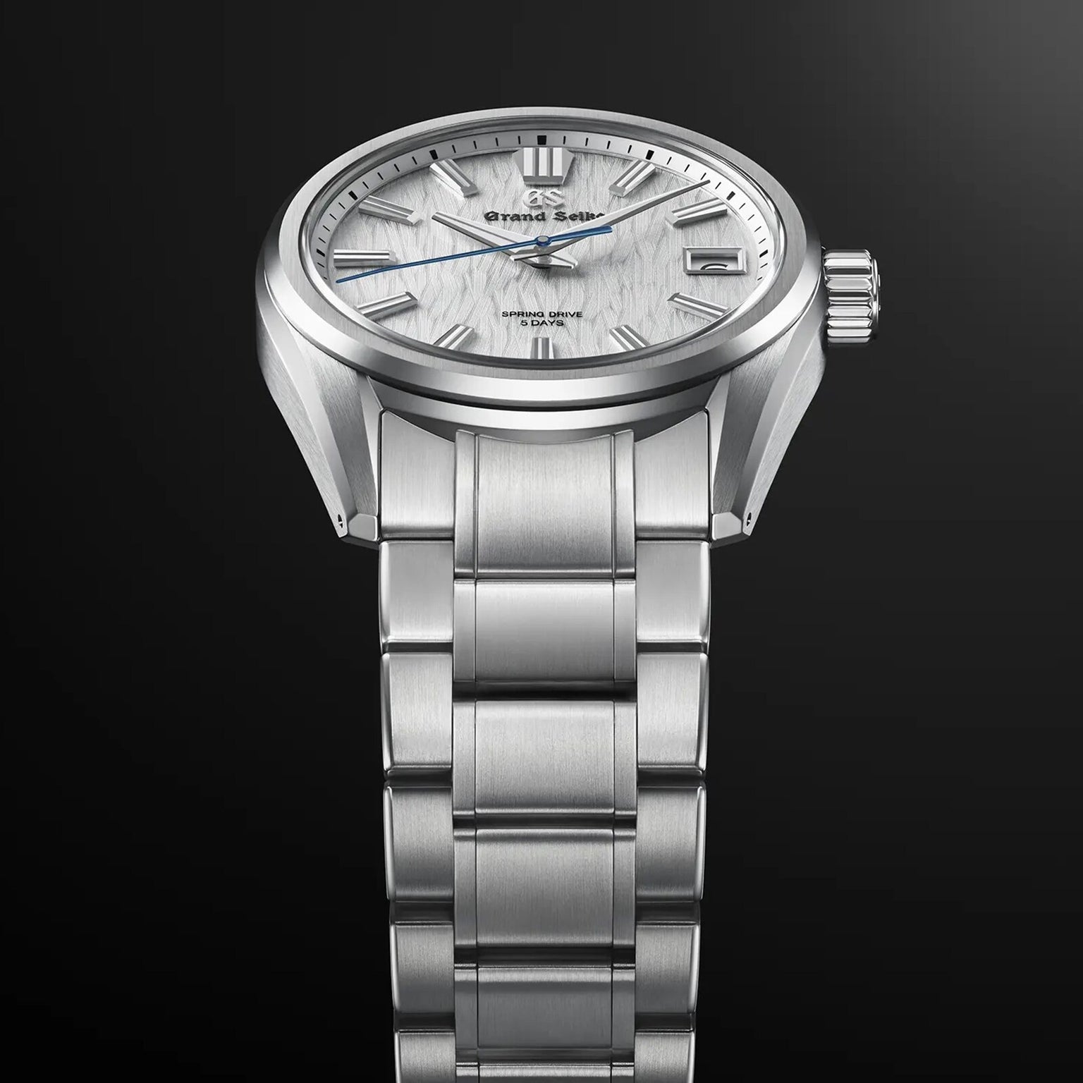 The Grand Seiko Spring Drive 5 Days Evolution 9, featuring a textured white dial and stainless steel bracelet, is elegantly displayed against a dark gradient background.