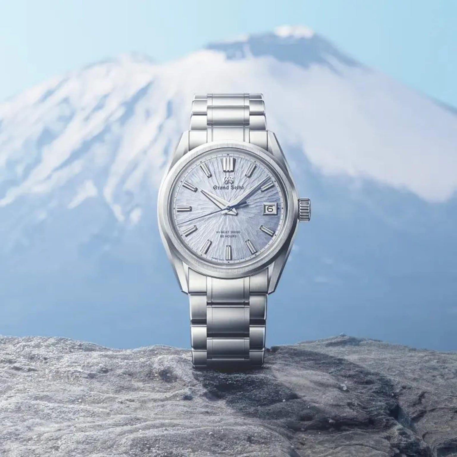 The Grand Seiko Hi-Beat 80 Hours Evolution 9 wristwatch with a textured silver face is displayed on a rock, set before the blurred backdrop of a snow-capped mountain landscape.