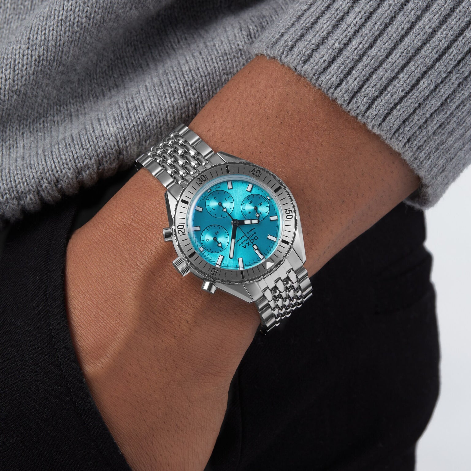 A person wearing the DOXA Sub 200 C-Graph II Aquamarine, a mechanical automatic chronograph with a turquoise dial and stainless steel bracelet, styled with a grey sweater and black pants.