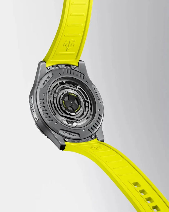 A close-up of the back of the Ulysse Nardin UR-FREAK wristwatch with a yellow strap, highlighting its detailed Swiss-made mechanical movement and engraved metal case against a light gray background.