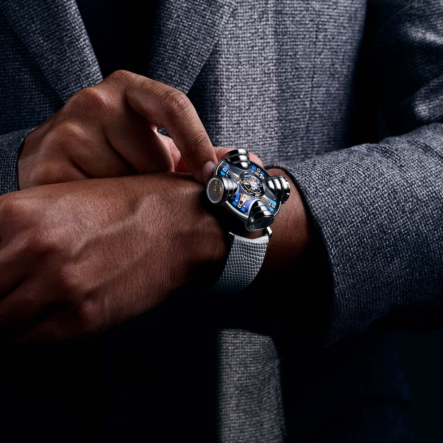 A person in a gray suit adjusts the MB&F HM11 Art Deco wristwatch, featuring a metallic band and skeletonised dial, highlighting its unique mechanical design on their left wrist.