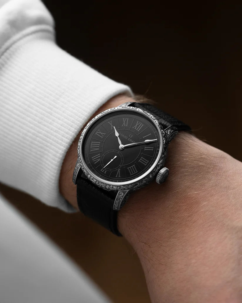 Black wristwatch with a dark face and Roman numeral markers on a wrist.