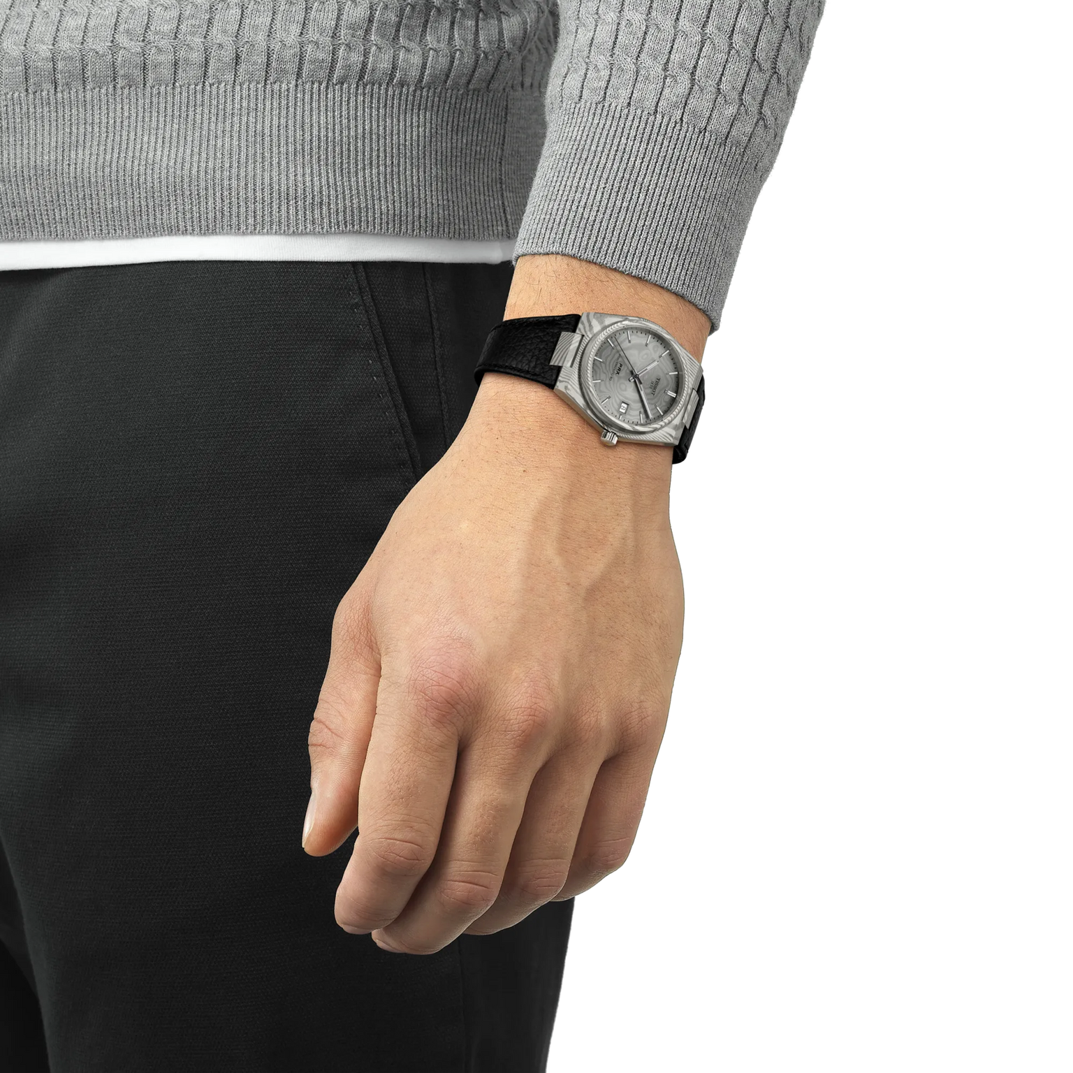 A person in a gray sweater and black pants stands with their left hand by their side, showcasing a Tissot PRX 38 watch with a black strap. The background is plain white.