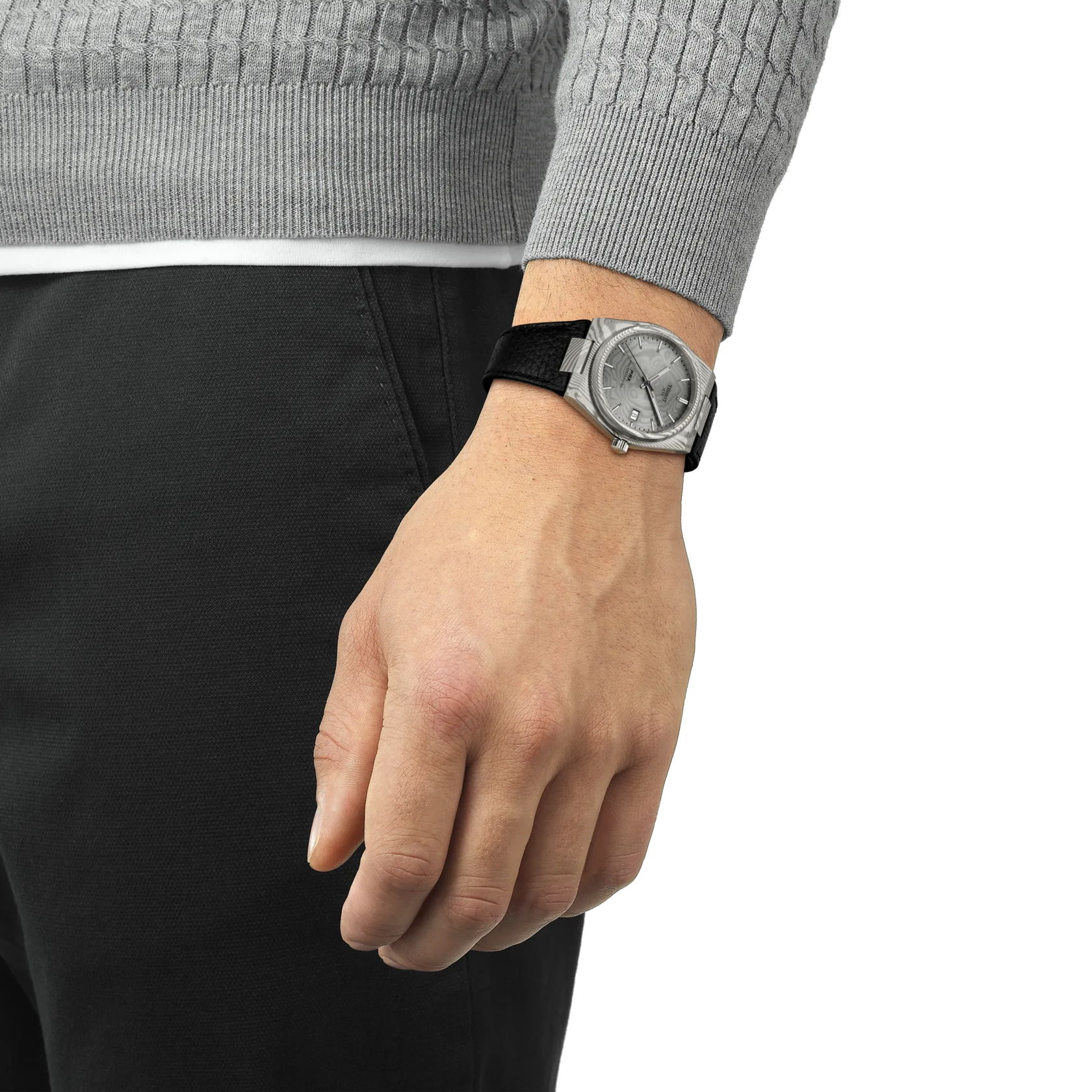 A person in a gray sweater and black pants stands with their left hand by their side, showcasing a Tissot PRX 38 watch with a black strap. The background is plain white.