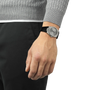 A person in a gray sweater and black pants stands with their left hand by their side, showcasing a Tissot PRX 38 watch with a black strap. The background is plain white.