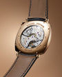 Rose gold square watch with exposed mechanical movement, blue leather strap, and Vacheron Constantin branding on dial.