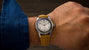 Oris watch with white dial, Arabic numerals, and tan suede strap on wrist wearing blue denim sleeve.