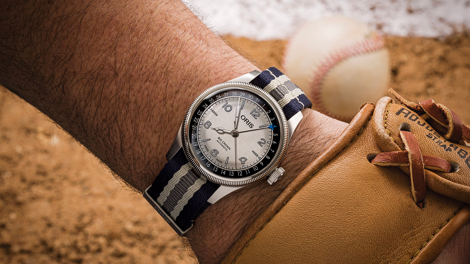 Stainless steel watch with white dial and navy striped fabric band worn on wrist next to baseball glove and baseball.