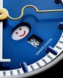 Blue watch dial featuring a smiling face design with red eyes and mouth, yellow hand, and "Swiss Made" text.