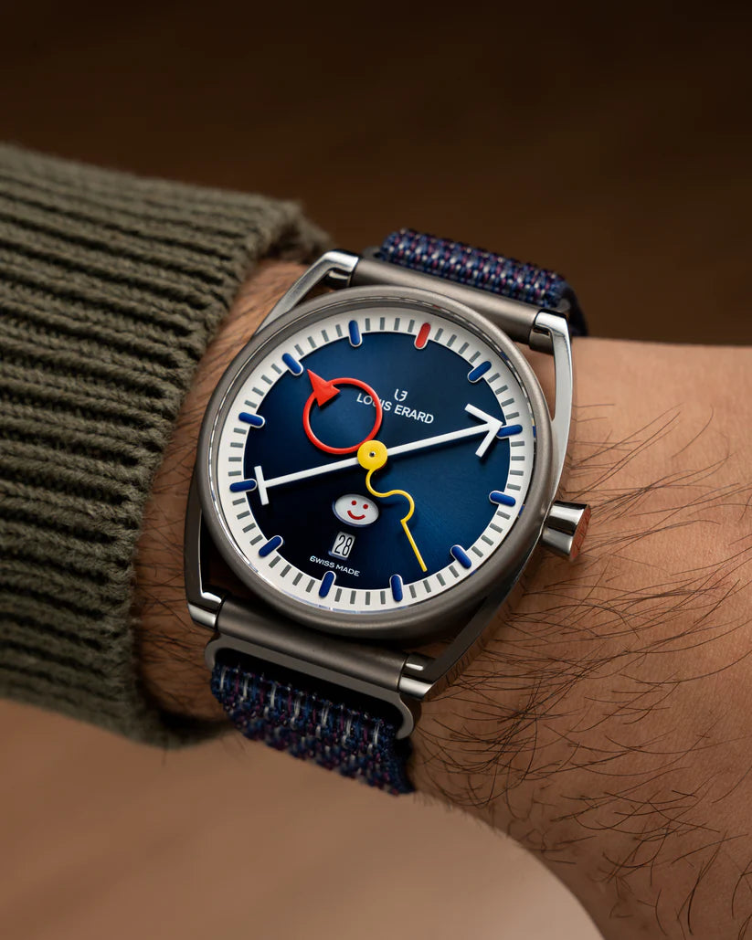 Stainless steel watch with blue dial, red moon hand, yellow center hand, smiley face subdial, and blue fabric strap worn on w