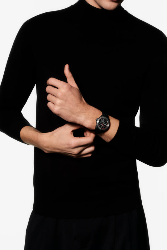 Person wearing a black turtleneck sweater and black pants with a watch on a white background