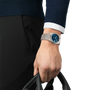 A person wearing a long-sleeve navy sweater and black pants holds a black bag. On their left wrist is a Tissot PRX 38 automatic watch with a blue face and silver case. The background is plain white.