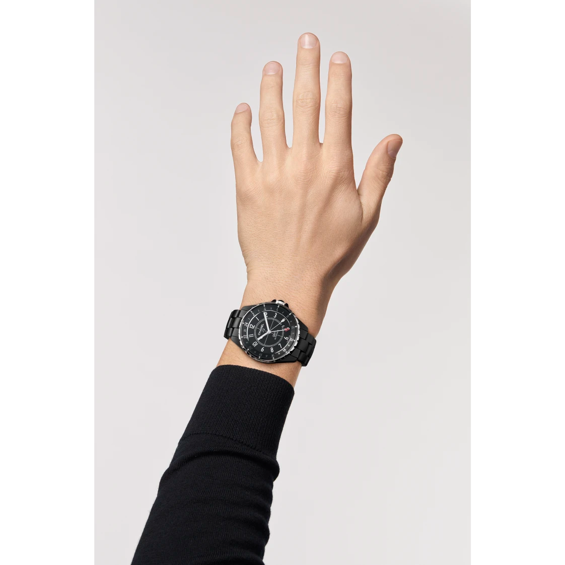 A raised left hand wears a Chanel J12 GMT wristwatch with a round face, high-precision white hour markers, and a black band. The wearer has on a long-sleeve black shirt against a plain, light-colored background.