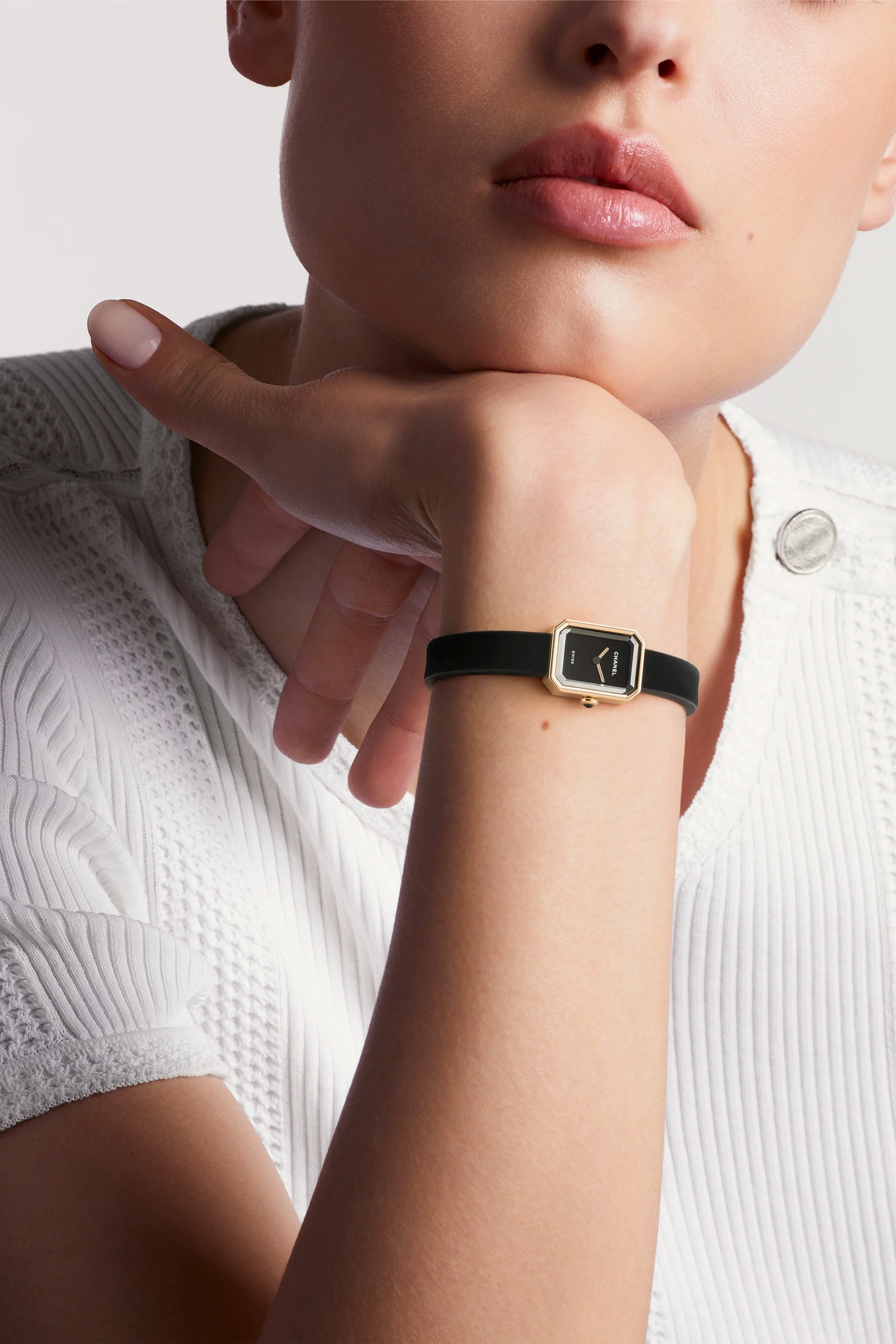 A close-up of a woman resting her chin on her hand, wearing a white textured top and the Chanel Première Ribbon watch with a rectangular black case, yellow gold frame, and black-lacquered dial. Her lips are slightly parted and her face is partially visible.