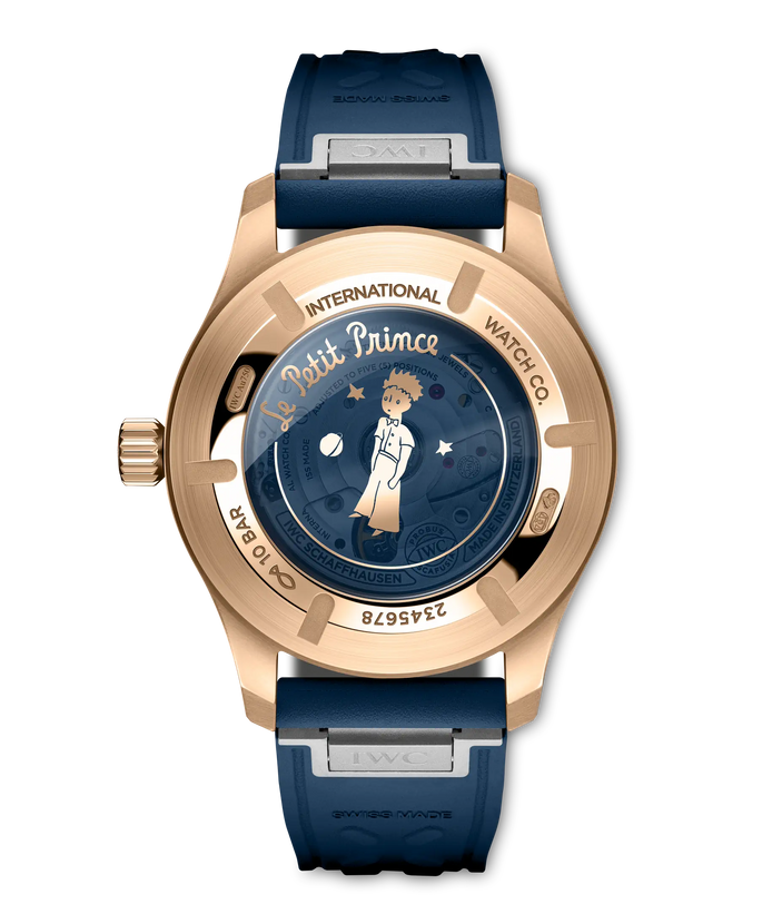 Rose gold pilot's watch with dark blue dial featuring Le Petit Prince illustration, blue rubber strap, and visible case back.
