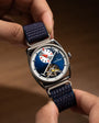 Square watch with blue dial, white hour markers, subsidiary dial, visible tourbillon, and dark blue fabric strap held in hand