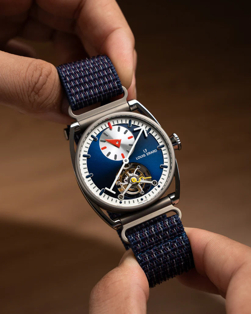 Square watch with blue dial, white hour markers, subsidiary dial, visible tourbillon, and dark blue fabric strap held in hand