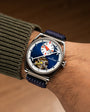 Stainless steel watch with blue dial, white subdial, red accents, visible tourbillon, and navy fabric strap worn on wrist.