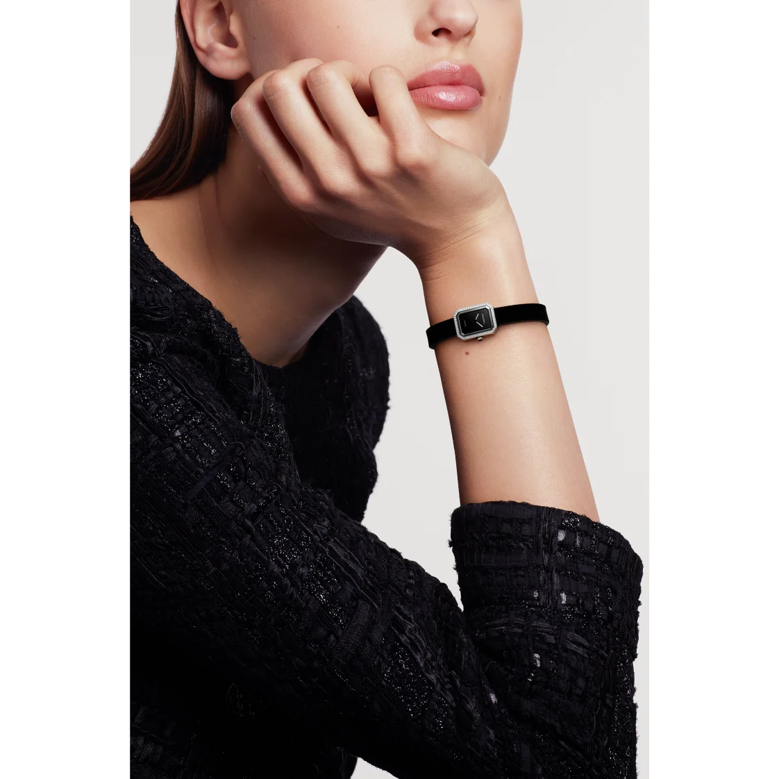 A woman in a textured black long-sleeve top rests her chin on her hand, showcasing the Chanel Première Ribbon—a small, square wristwatch with a black-lacquered dial and black strap—against a plain white background.