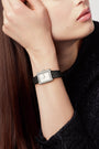 A woman with straight brown hair, wearing a textured black top, rests her chin on her hand and shows off the Chanel Boy·Friend Small Model watch with a square face and black calfskin strap. She has a neutral expression.