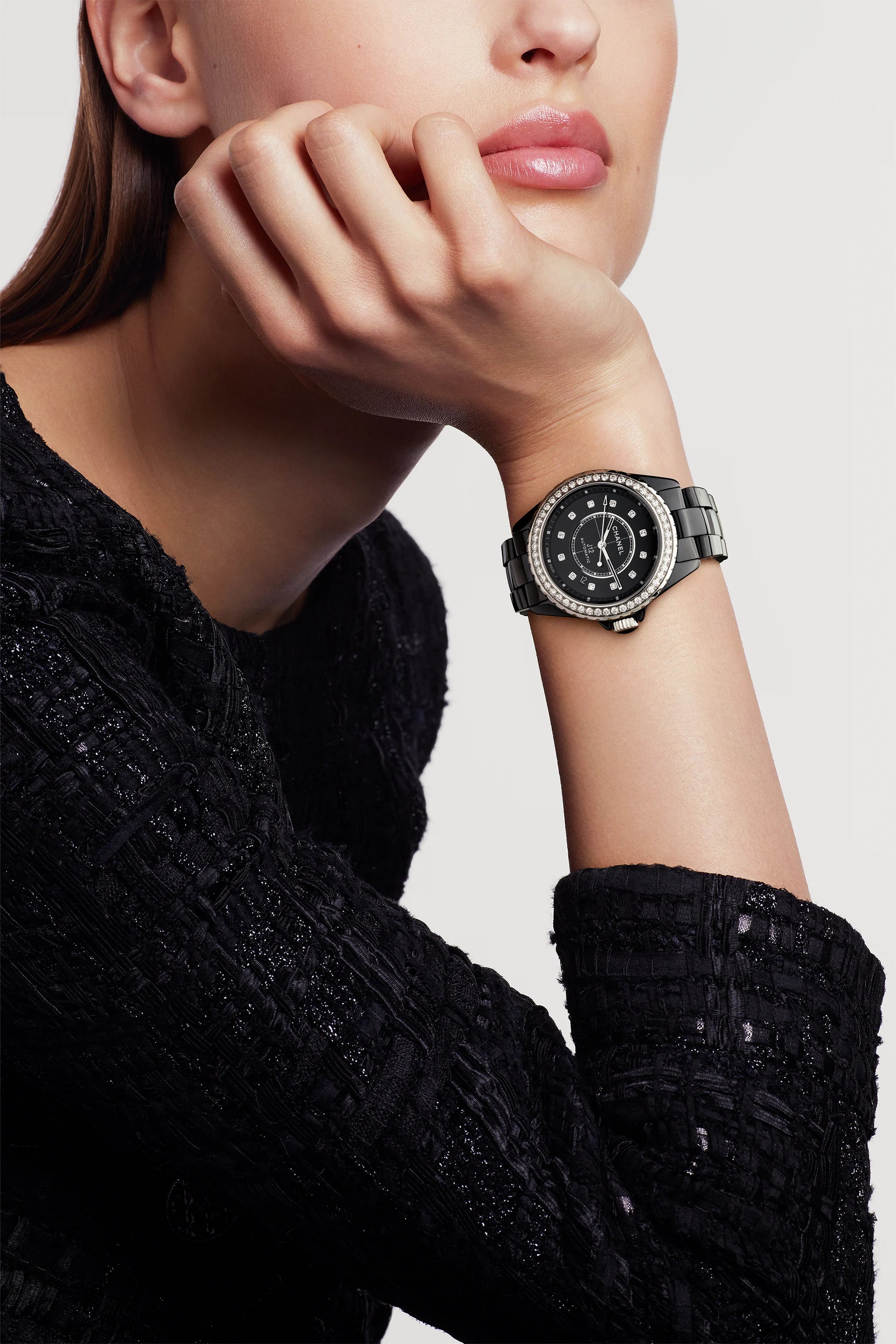 A woman in a textured black top rests her chin on her hand, showcasing the Chanel J12 Diamond Bezel—a highly resistant ceramic and steel wristwatch with a diamond bezel and an elegant, modern design.