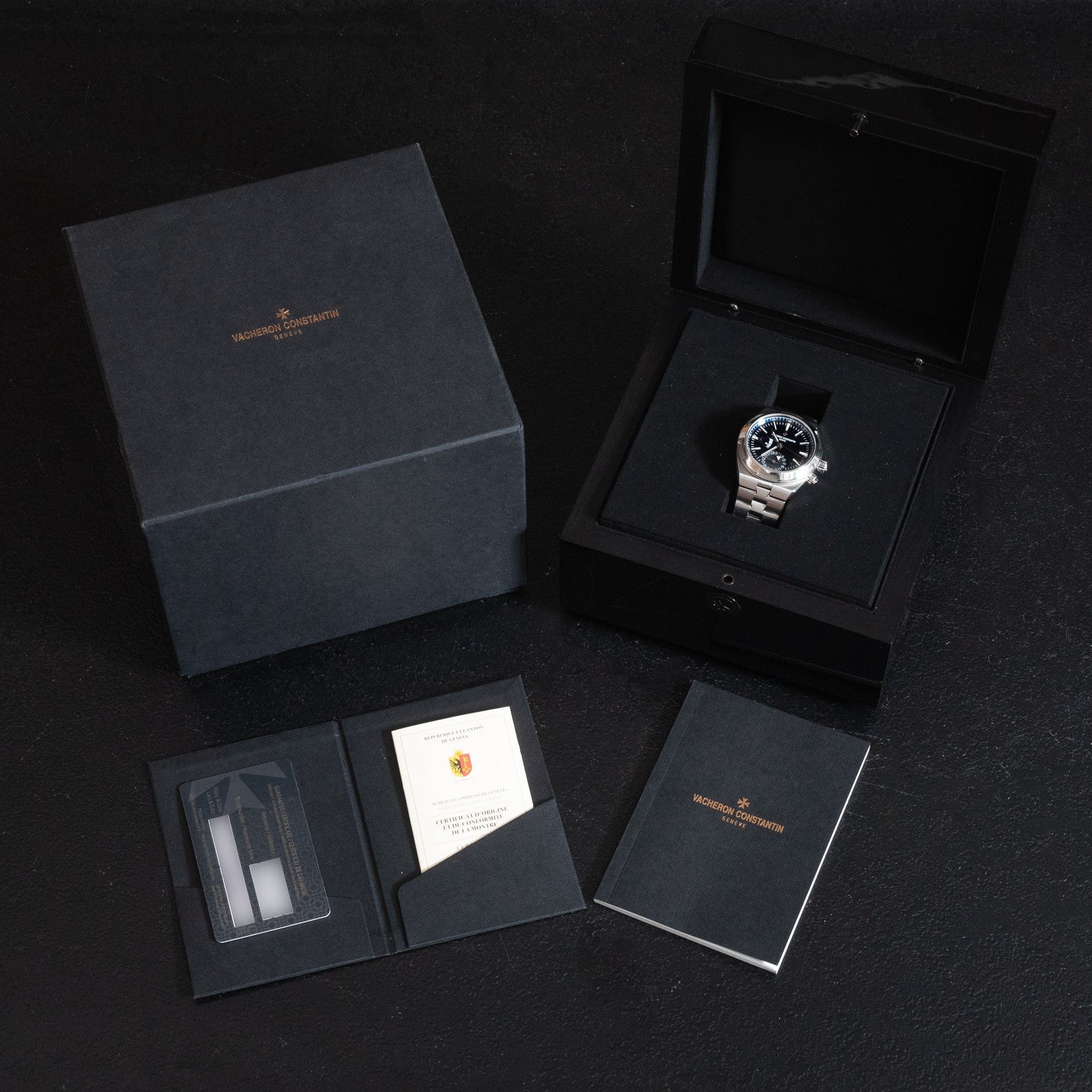 The Vacheron Constantin Overseas Dual-Time luxury watch is presented in its original black box with warranty cards, booklet, and packaging, all elegantly displayed on a dark surface.