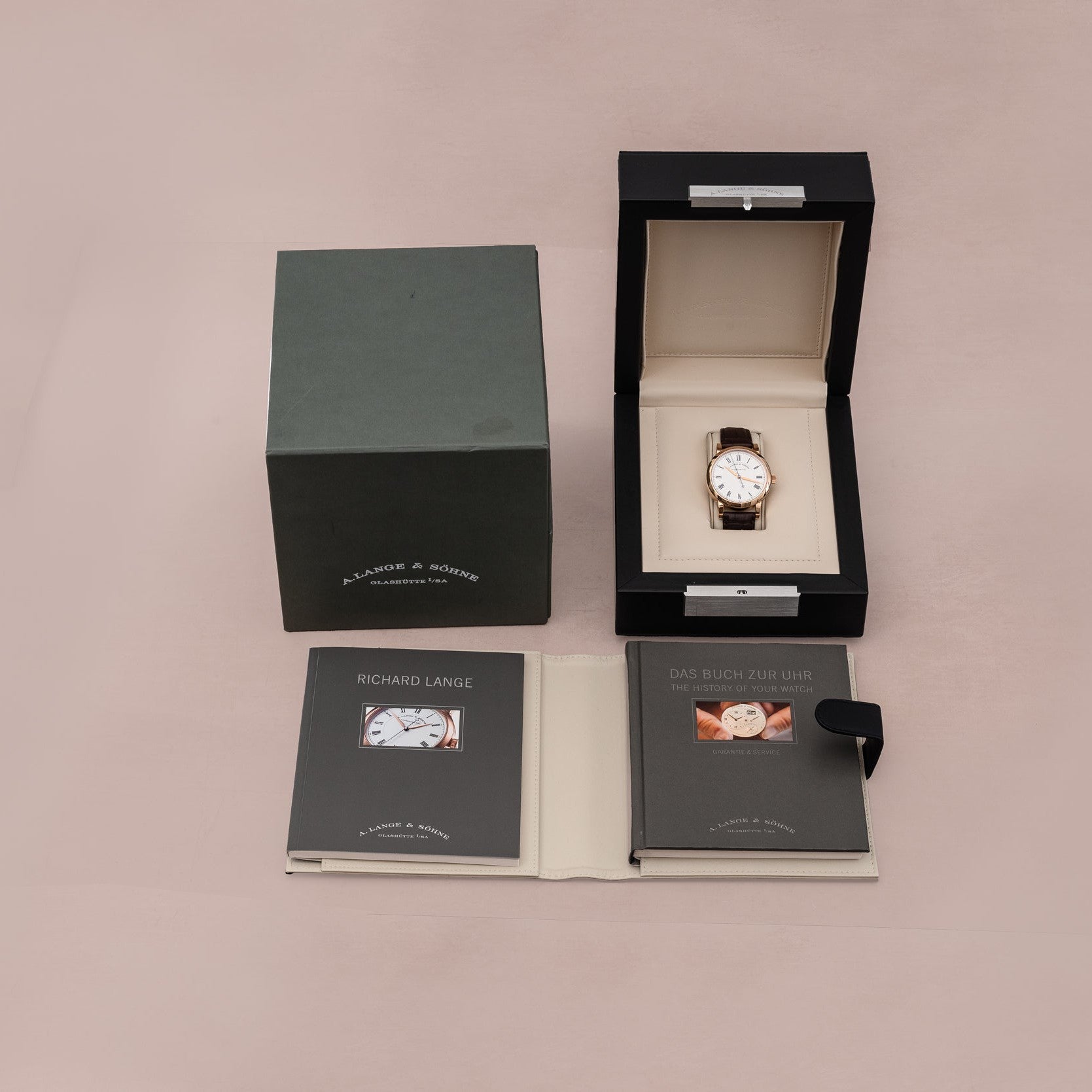 A Lange & Sohne Richard Lange luxury wristwatch in an open box with a green case and two booklets, displayed on a light pink surface.