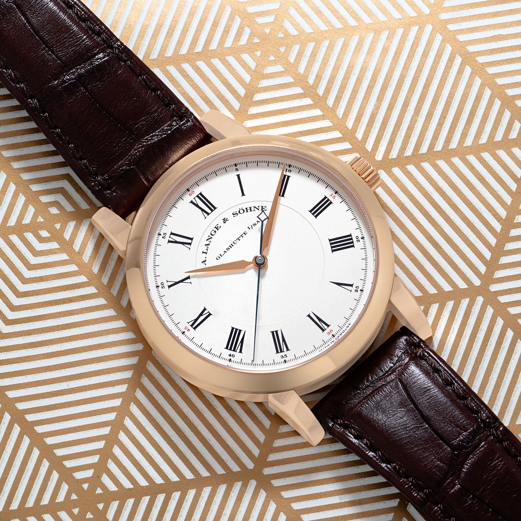 The Richard Lange by Lange & Sohne features a white face, black Roman numerals, and a brown leather strap, displayed on a geometric gold background.
