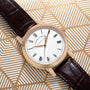 The Richard Lange by Lange & Sohne features a white face, black Roman numerals, and a brown leather strap, displayed on a geometric gold background.