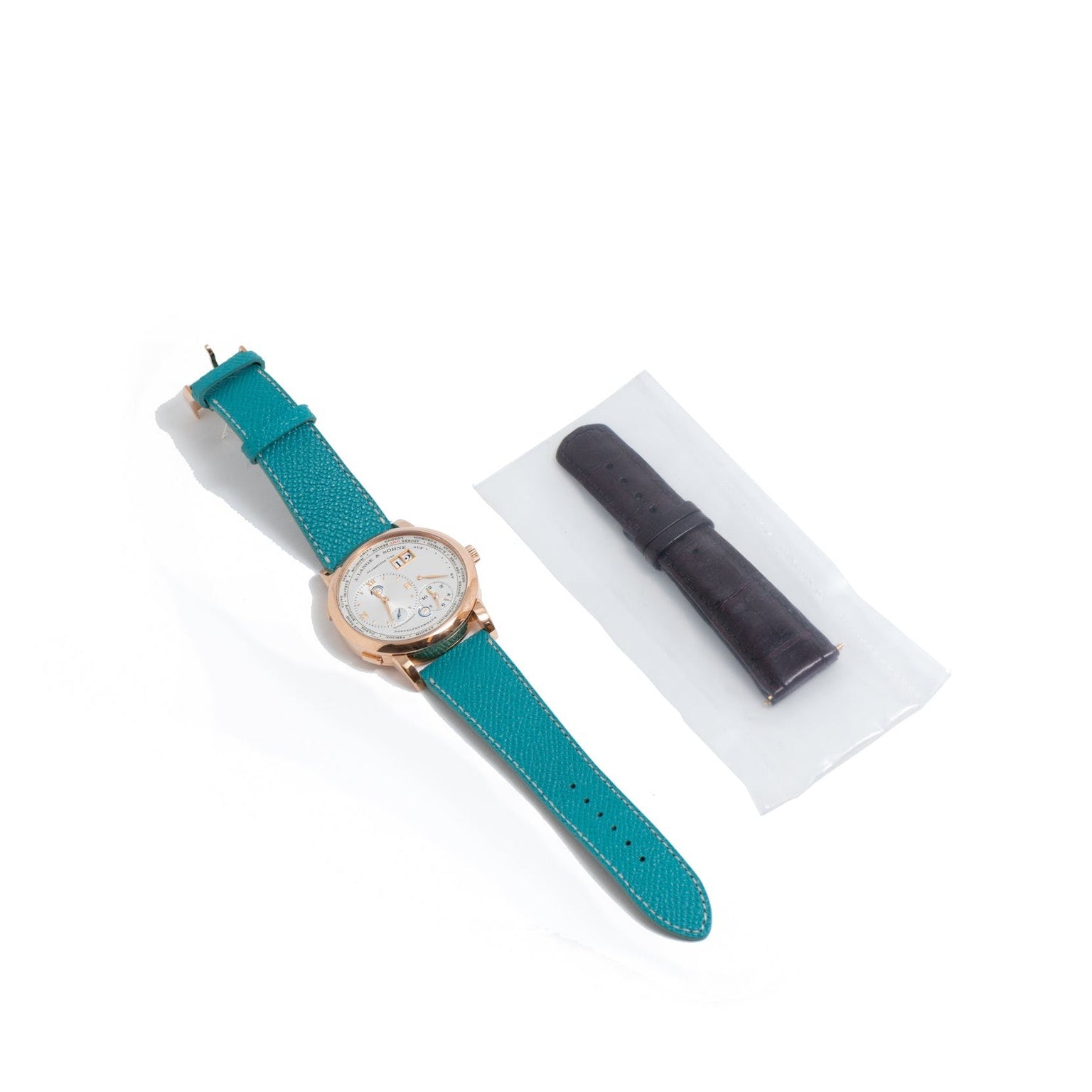 A Lange & Sohne Lange 1 Timezone wristwatch with a teal strap sits beside a packaged dark brown replacement strap, displayed on a white background.