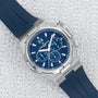 The Vacheron Constantin Overseas Chronograph is a luxury silver watch with a blue face and blue rubber strap, shown on a textured light surface.
