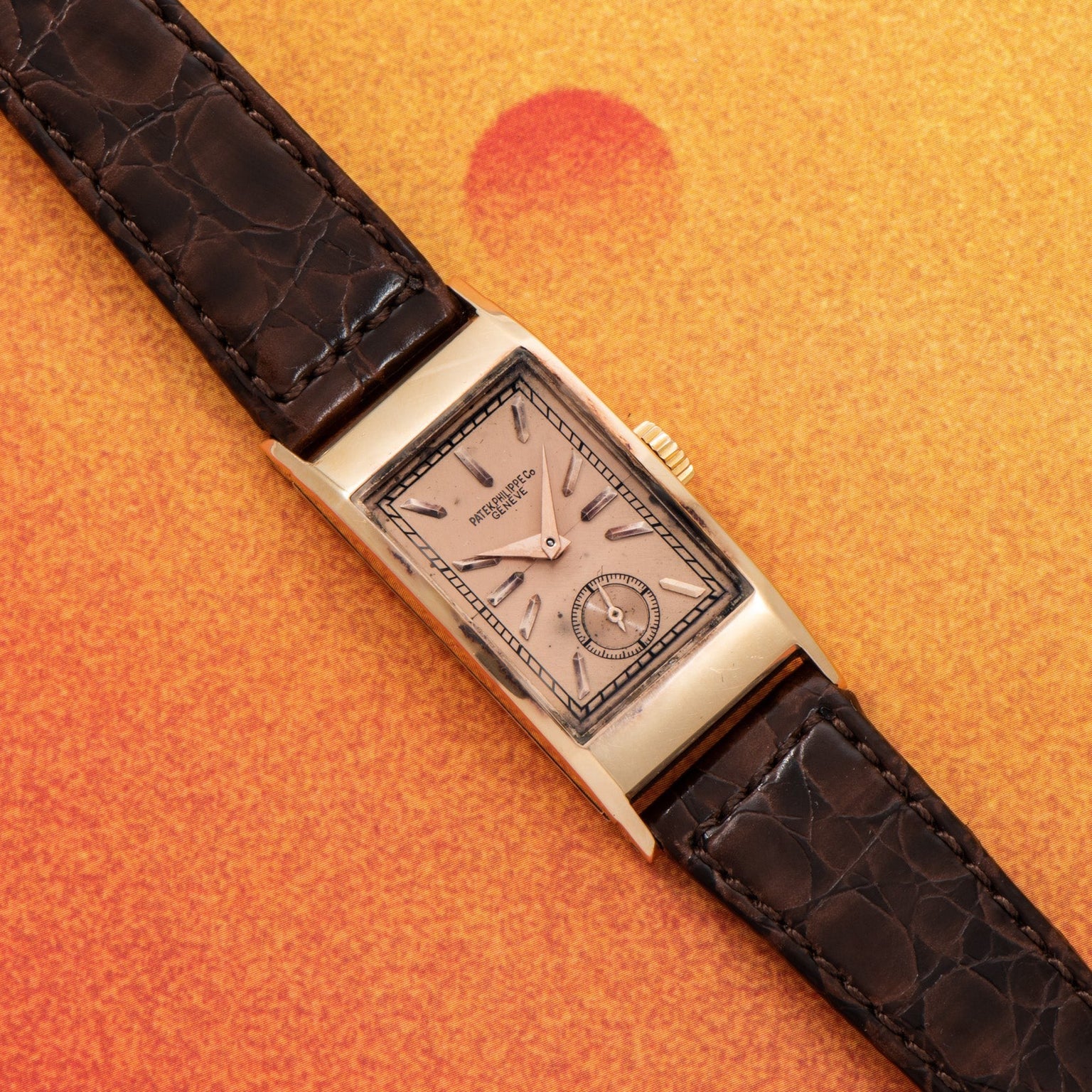 The Patek Philippe Tegolino Dress Watch, featuring a rectangular gold case and dark leather strap, is displayed on an orange textured background.