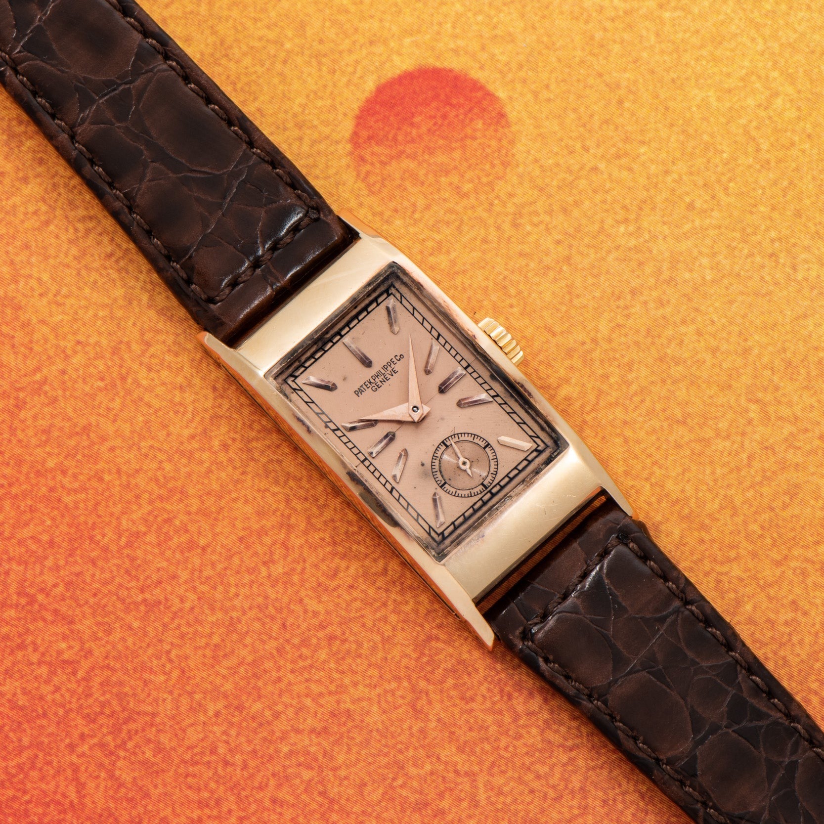 The Patek Philippe Tegolino Dress Watch, featuring a rectangular gold case and dark leather strap, is displayed on an orange textured background.