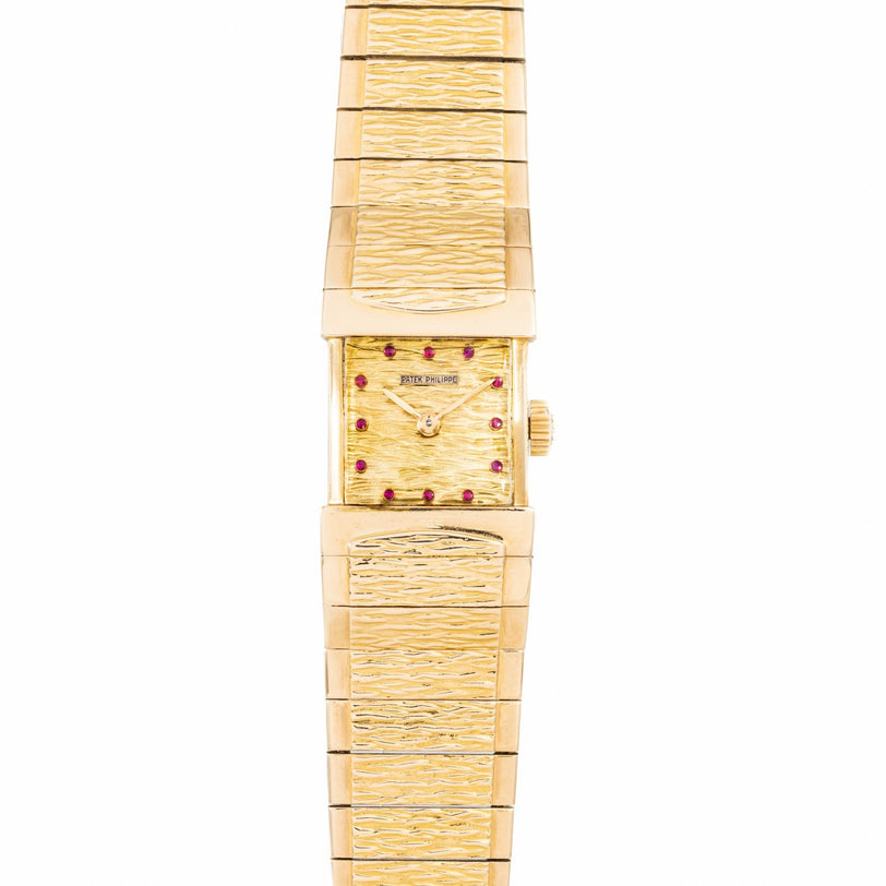 The Patek Philippe Yellow Gold Integrated Dress Watch features a rectangular case, textured band and dial, red gem hour markers, and a simple gold crown.