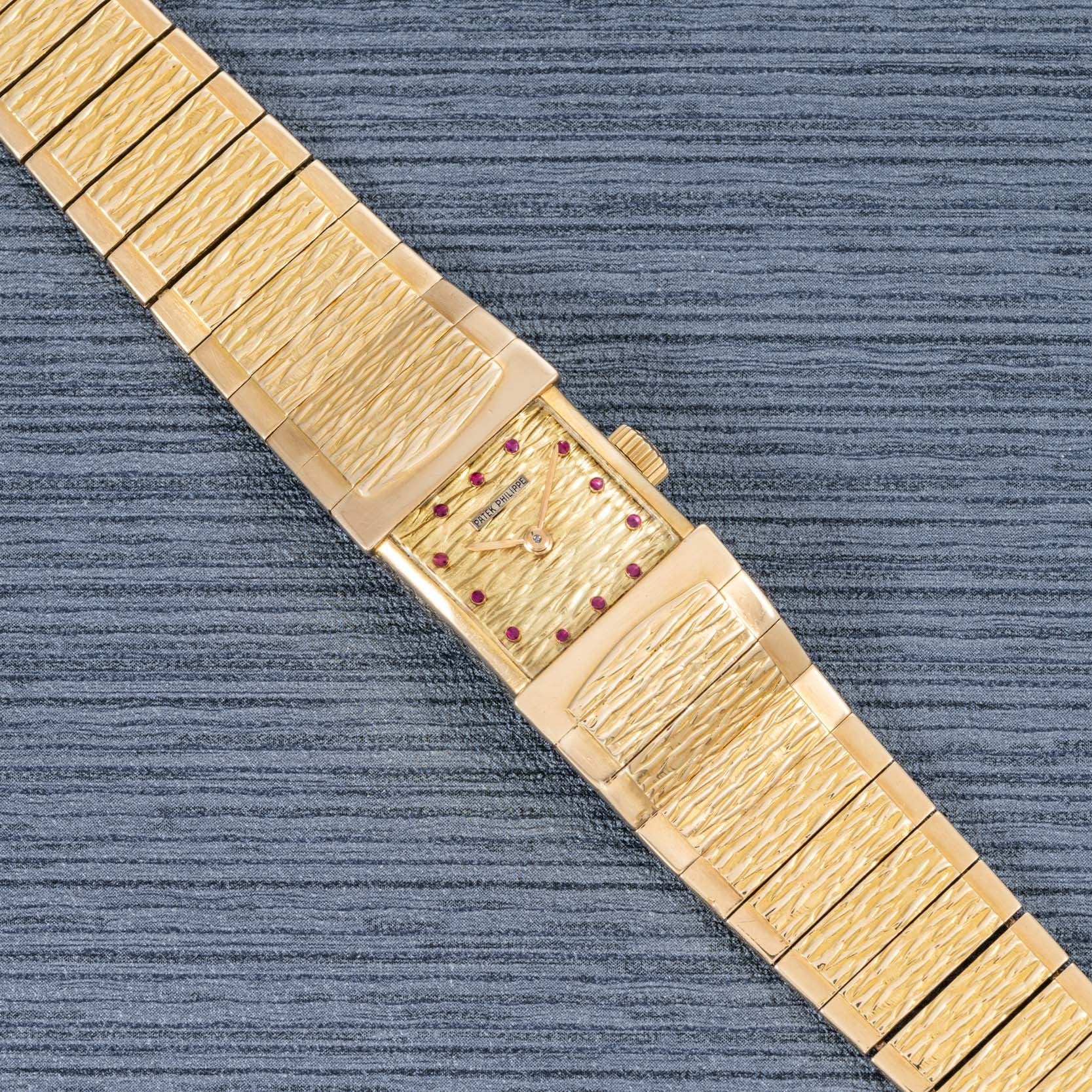 The Patek Philippe Yellow Gold Integrated Dress Watch features a textured band, rectangular face, and ruby hour markers, displayed on a gray textured surface.