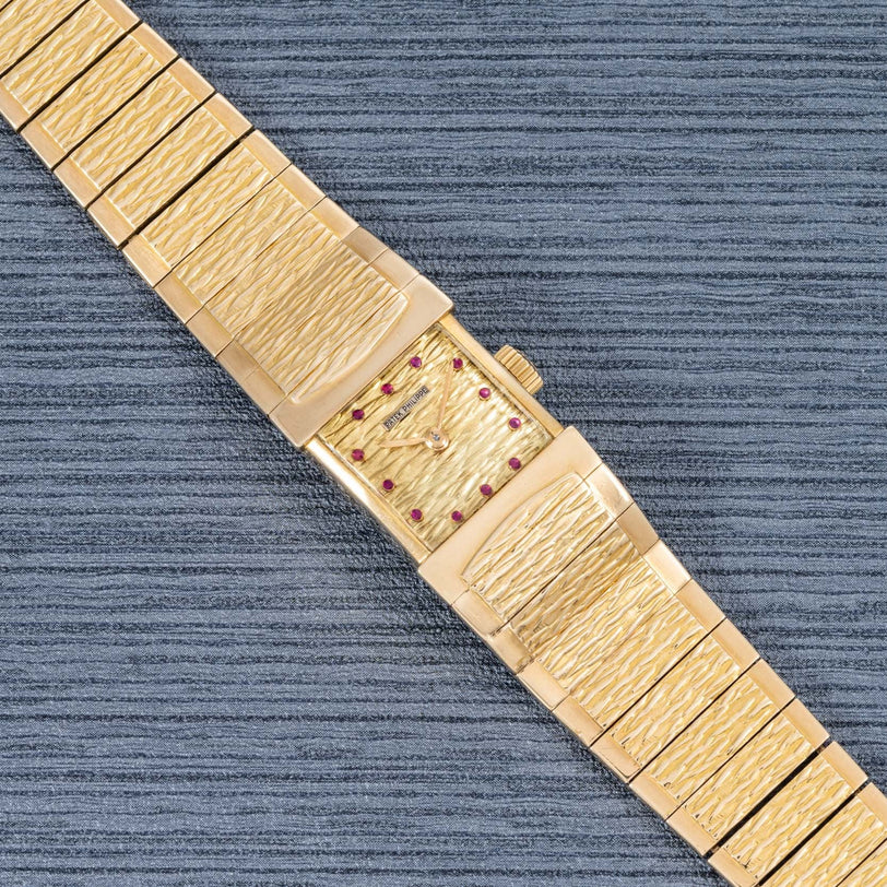 The Patek Philippe Yellow Gold Integrated Dress Watch features a textured band, rectangular face, and ruby hour markers, displayed on a gray textured surface.