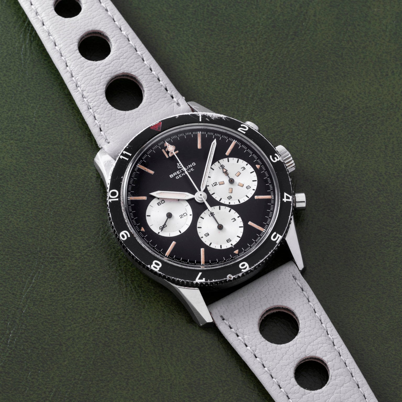 A Breitling Co-Pilot Chronograph wristwatch in black and silver features a perforated gray leather strap, displayed against a green background.