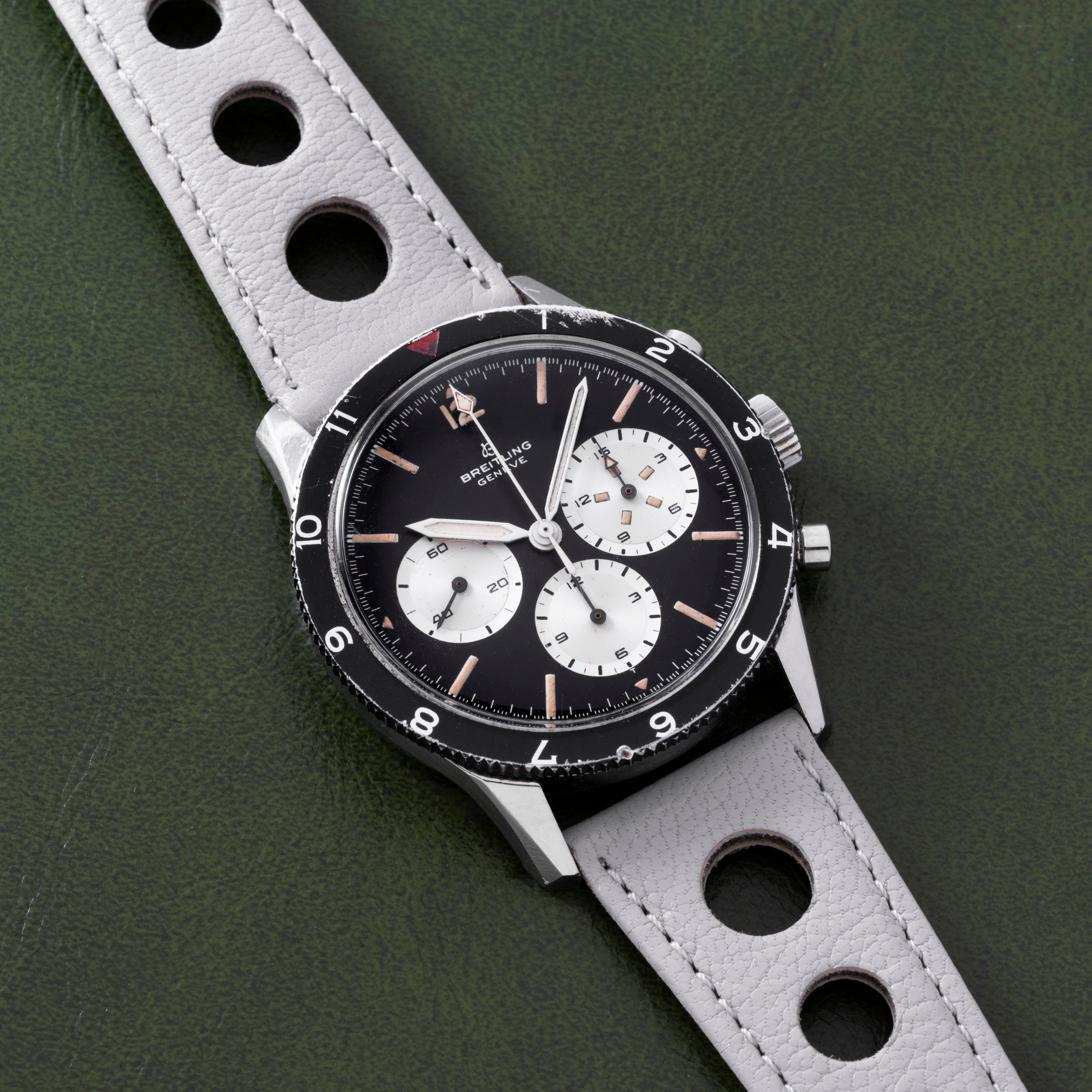 A Breitling Co-Pilot Chronograph wristwatch in black and silver features a perforated gray leather strap, displayed against a green background.