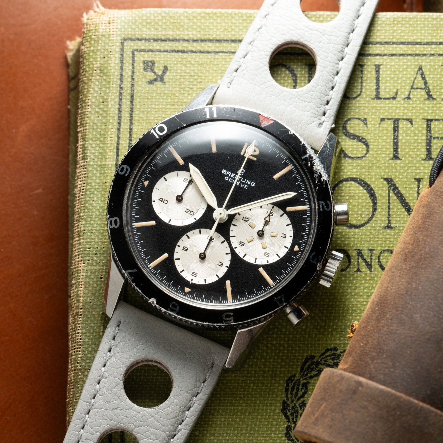 A Breitling Co-Pilot Chronograph with a white perforated strap rests on a green book.