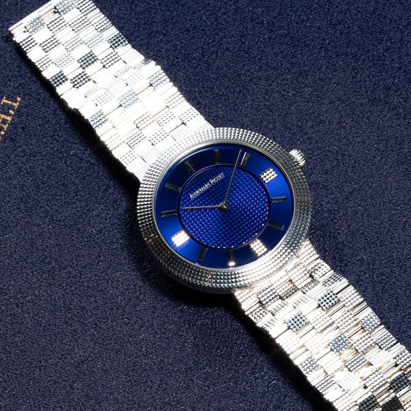 Audemars Piguets Classic White Gold Ultra-Thin watch, featuring a sleek metallic strap, rests elegantly on a dark textured surface.