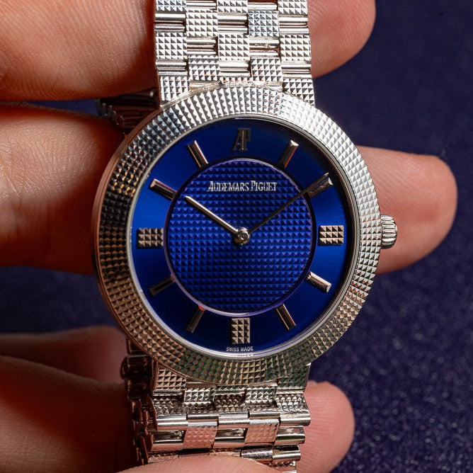 A hand holds a silver Audemars Piguet Classic White Gold Ultra-Thin wristwatch with a blue textured dial and silver hour markers.